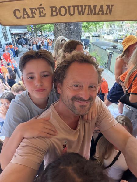 A man and a boy take a selfie on a bustling street, likely Prinsengracht, Centrum, Amsterdam, with a large crowd of people dressed in orange visible in the background